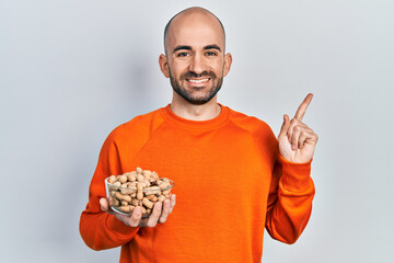 Young bald man holding peanuts smiling happy pointing with hand and finger to the side