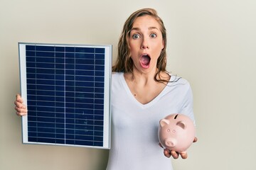 Young blonde woman holding photovoltaic solar panel and piggy bank afraid and shocked with surprise...