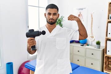Young indian physiotherapist holding therapy massage gun at wellness center strong person showing arm muscle, confident and proud of power