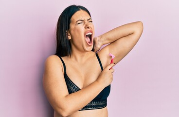 Beautiful brunette woman shaving armpit hair with razor angry and mad screaming frustrated and furious, shouting with anger. rage and aggressive concept. - obrazy, fototapety, plakaty
