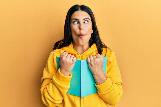 Beautiful brunette woman holding book making fish face with mouth and squinting eyes, crazy and comical.