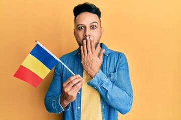 Young arab man holding romania flag covering mouth with hand, shocked and afraid for mistake....