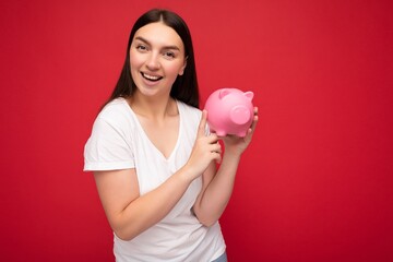 Fototapeta na wymiar Photo of happy positive smiling adult beautiful attractive brunette woman with sincere emotions wearing casual white t-shirt isolated over red background with copy space and holding pink pig savings
