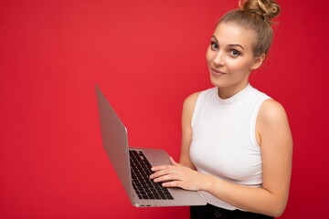 close-up shot of beautiful asking blond young girl student with gathered hair wearing white t-shirt holding computer laptop typing on keyboard looking at camera isolated over red wall background