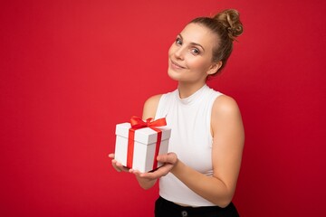 Pretty happy young blonde female person isolated over red background wall wearing white top holding gift box and looking at camera