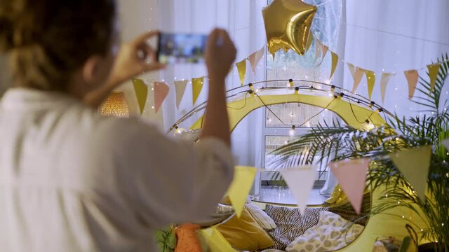 Woman takes picture with phone of stylish children room with bright play tent decorated for birthday celebration in light spacious room close view