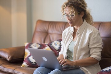 Profile of smiling woman in eyeglasses and curly hair watching media content using laptop on comfortable leather sofa at home. Happy woman working on laptop at home