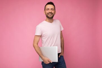 Handsome smiling and happy man holding laptop computer looking at camera in t-shirt on isolated pink background