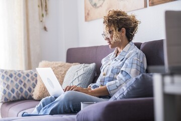 Beautiful young woman in eyeglasses working on laptop sitting on sofa. Businesswoman working from home. Young woman in casuals sitting on couch. Relaxed woman using laptop on couch at home