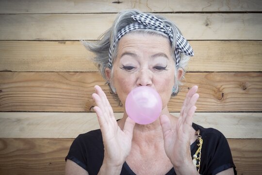 Portrait Of Senior Woman In T Shirt And Bandanna Showing Balloon Of Bubble Gum. Elderly Woman Having Fun Against Wooden Wall. Old Playful Woman Blowing Bubble Gum