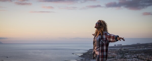 Panoramic view of young woman in sunglasses standing with arms stretched during sunset against cityscape and sea. Beautiful woman standing at sea coast. Carefree woman standing outdoors by seascape