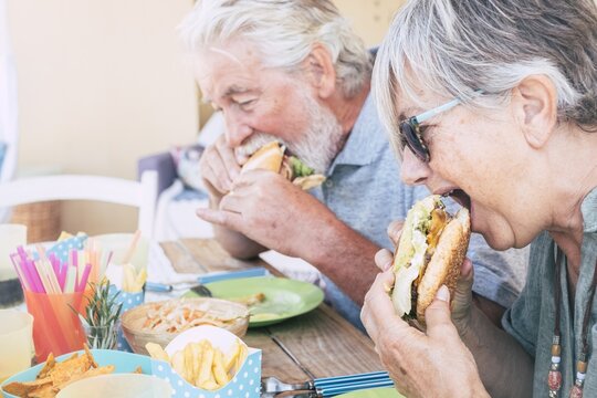 Senior Couple Taking A Bite Of Burger At Restaurant. Couple Having Lunch At Cafe. Old Couple With Gray Hair Eating Sandwich At Outdoor Restaurant During Summer Holiday