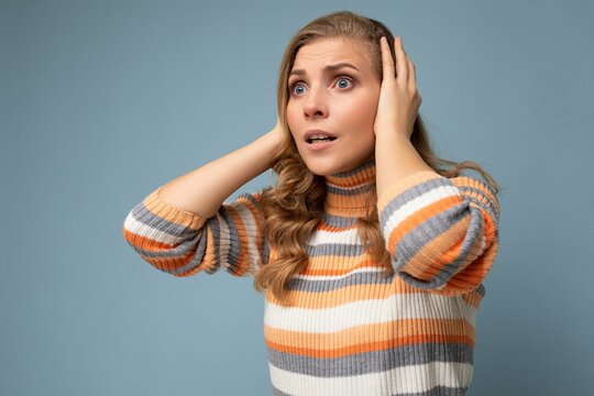 Young shocked pretty nice blonde curly woman with sincere emotions wearing casual striped pullover isolated on blue background with free space and covering ears trying not to hear