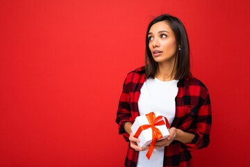 Fototapeta na wymiar Photo shot of pretty positive thoughtful young brunette woman isolated over red background wall wearing white casual t-shirt and red and black shirt holding white gift box with red ribbon and looking