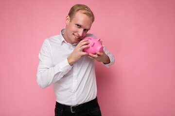 Photo of happy smiling young handsome blond man with sincere emotions wearing casual white shirt isolated over pink background with empty space and holding pink pig penny bank. Money box concept