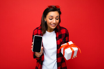 Shot of beautiful positive amazed young brunette woman isolated over red background wall wearing white casual t-shirt and red and black shirt holding white gift box with red ribbon and mobile phone