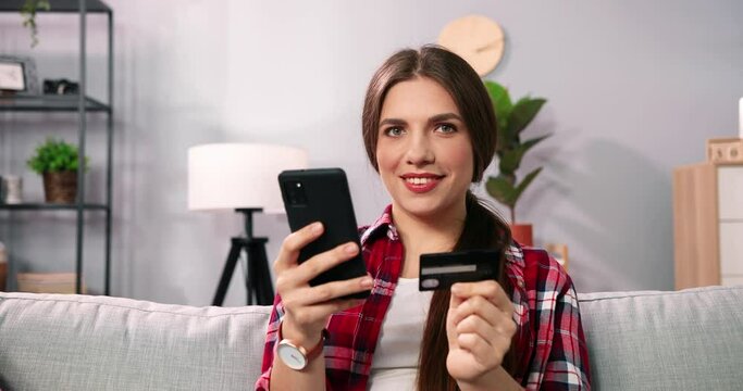 Dreaming about great shopping. Beautiful caucasian woman sitting on sofa ine room and using smartphone while making shopping online payment. Modern woman private time at home and internet shopping