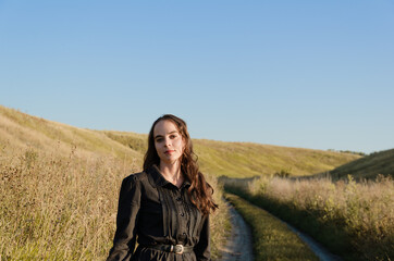 Naklejka na ściany i meble Young brunette woman looking at camera wearing black dress standing outdoors between hills during sunset, autumn season.