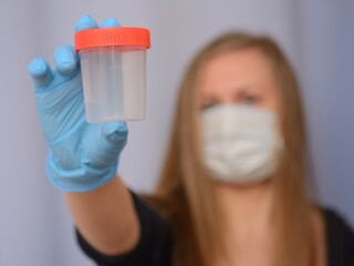 Hand in medical gloves holding a empty jar for analysis in focus, a doctor in a mask in a hospital room in blur.