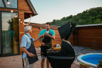 Two smiling men (young and old) drinking beer in the summer garden while barbecuing