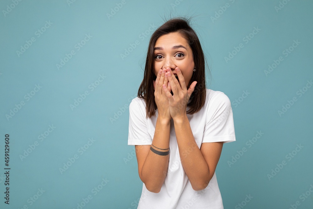 Wall mural Portrait of positive surprised young beautiful brunette woman with sincere emotions wearing casual white t-shirt for mockup isolated on blue background with copy space and covering mouth with hands