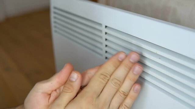 Man warms his frozen hands near convector electric heater at home.