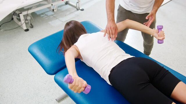 Overhead shot of a Caucasian woman lying on her abdomen, doing shoulder stabilization exercises with small weights, with a help of the male physiotherapist