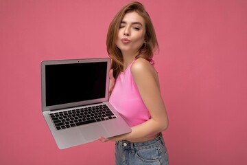 Close-up photo of Beautiful young blond woman holding computer laptop wearing pink crop top looking at camera with close eyes and giving a kiss isolated on pink background