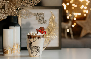 A sign with the inscription Happy New Year, a mug with hot cocoa, marshmallows and a gingerbread man on the table in the home interior of the living room. A cozy concept of the Christmas and winter.