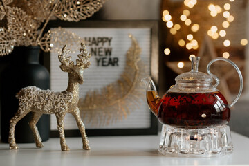 A sign with the inscription Happy New Year, a deer, a golden bouquet on the table and a teapot with tea on the table in the home interior of the living room. A cozy concept of a holiday and winter.