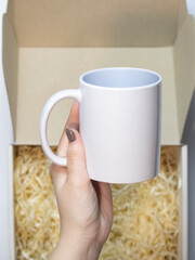 female hands with manicure, holding a white sublimation mug on a blurred background of a box with wood shavings. cup mockup