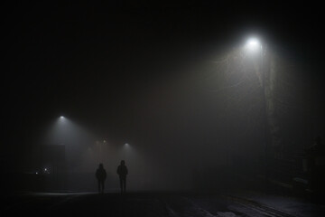 Two mysterious hooded men walk away down a dark foggy road, dimly lit