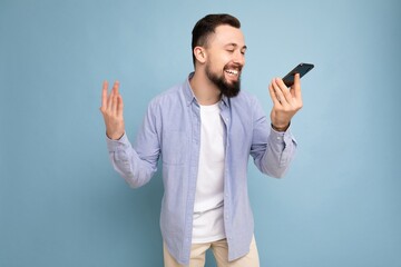 Photo shot of happy good looking young brunet bearded man wearing casual blue shirt and white t-shirt poising isolated on blue background with empty space holding in hand and communicating talking on