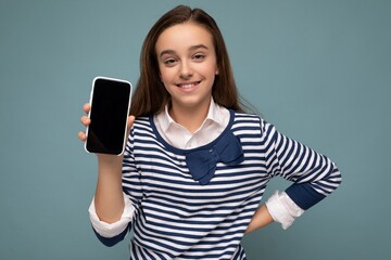 Fototapeta premium Pretty positive smiling girl wearing striped longsleeve standing isolated on blue background with copy space holding smartphone showing phone in hand with empty screen display for mockup pointing at