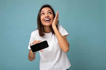Naklejka premium Photo of pretty positive amazed young brunette woman good looking wearing white t-shirt standing isolated on blue background with copy space holding smartphone showing phone in hand with empty screen