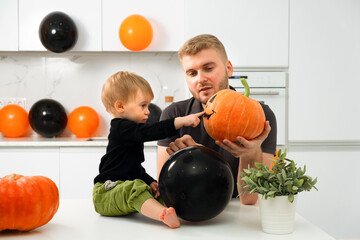 Fototapeta premium Handsome caucasian blond man show pumpkin horror face to 1 year old infant in home decoreted kitchen,modern interior.Happy Halloween day with family.Father,kid spend time,celebrate holiday together