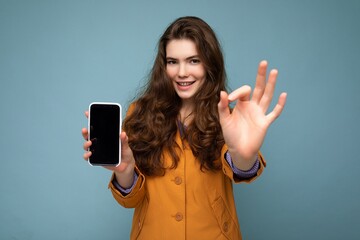 Beautiful young brunette woman wearing orange jacket isolated over blue background holding in hand and showing mobile phone with empty display for mockup looking at camera and showing okay gesture