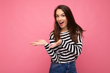 Portrait photo of young happy positive beautiful brunette woman with sincere emotions wearing casual striped pullover isolated on pink background with copy space and pointing hands at free space for