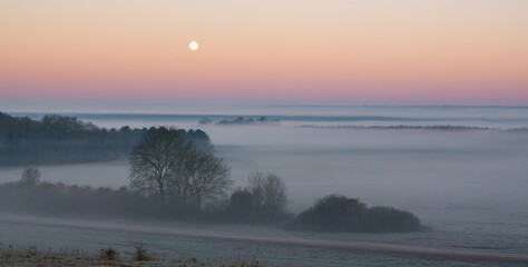 Obraz na płótnie Canvas an early morning mist filled valley with the snow moon low in the sunrise sky 