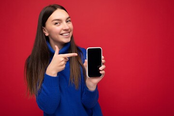 Photo of pretty happy young brunette woman wearing casual blue sweater isolated over red background with empty space holding in hand mobile phone and showing smartphone with empty screen for mockup