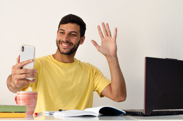 A positive young man in a yellow tshirt makes a video call on a smartphone on a background of a white wall, looks into the camera of the gadget and greets with his palm. 