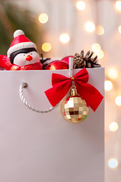 A white paper bag with a gold ball, a red bow with a Christmas tree branch and a toy penguin in a Santa's hat inside on a background of blurred bokeh lights. New Year's gift. Mockup for text, design