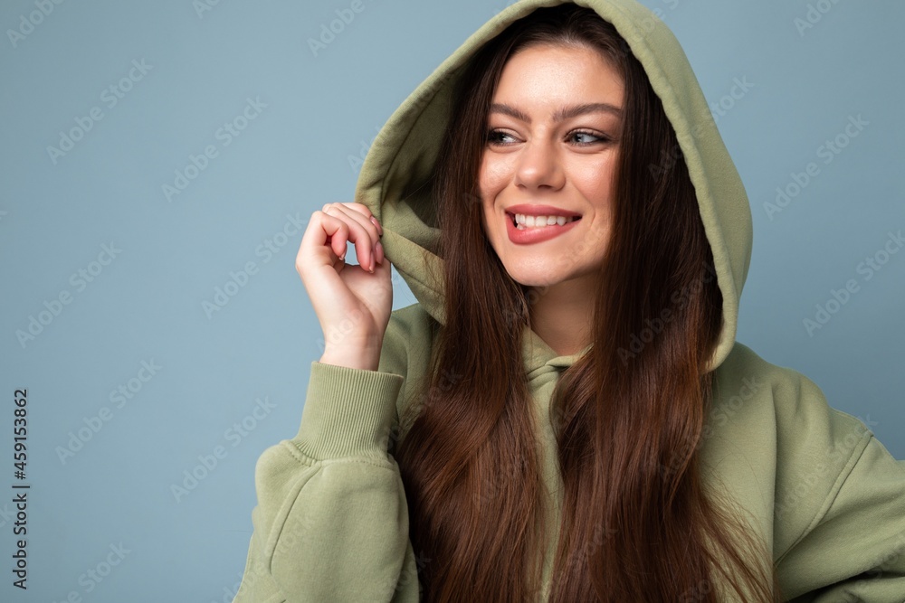 Wall mural Portrait of young beautiful smiling girl in stylish hipster green hoodie. Sexy carefree woman posing near blue wall. Positive model having fun - Wall murals