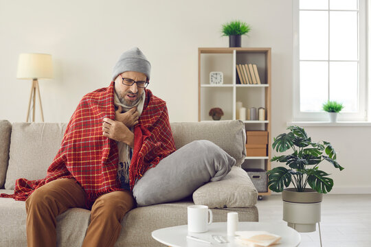 Unhappy Man All Wrapped In Warm Clothes Suffering From Symptoms Of Common Cold Or Flu. Sad Sick Middle Aged Man Who Has Sore Throat And Fever Sitting On Sofa At Home And Coughing