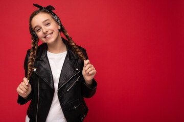 Photo shot of pretty positive smiling brunette little female teenager with pigtails wearing stylish black leather jacket and white t-shirt standing isolated over red background wall looking at camera