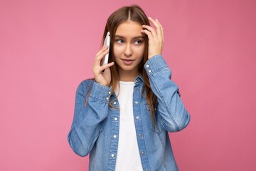 Fototapeta premium Photo of beautiful sexy thinking young blonde woman wearing casual blue jean shirt isolated over pink background holding in hand and talking on mobile phone looking down to the side