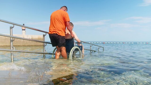 Handheld shot of a young Caucasian man in a wheelchair slowly pushed back, along the ramp, out of seawater on the beach.