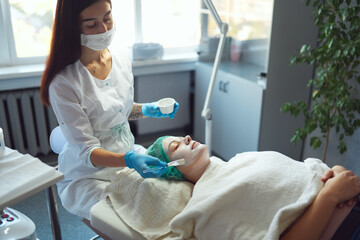 Beautician covering woman facial skin with moisturizing cleansing mask during skin care procedure in a beauty salon.