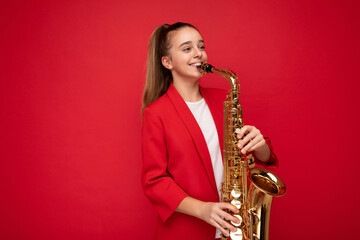 Fototapeta na wymiar Shot of pretty happy smiling brunette little girl wearing stylish red jacket standing isolated over red background wall playing saxophone looking to the side