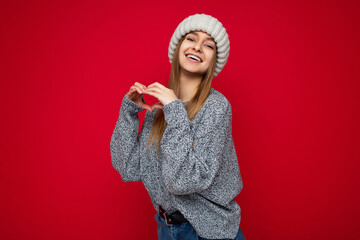 Portrait of positive smiling young beautiful dark blonde woman with sincere emotions wearing grey sweater and beige knitted hat isolated over red background with free space and showing heart shape